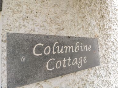 A close-up of a dark rectangular sign with the words Columbine Cottage on a textured white wall at Columbine Cottage in Hawkshead