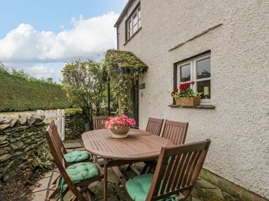 An outdoor patio with a wooden table and six chairs with green cushions next to a white wall with a window and flowers at Columbine Cottage in Hawkshead