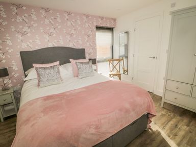 A bedroom with a double bed with pink bedding wallpaper with floral design a wooden chair a mirror a white door and a wardrobe at Forget Me Not Lodge Bamburgh