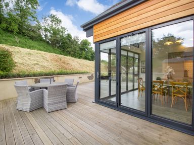 An outdoor wooden deck with a round table and six wicker chairs next to a building with large glass sliding doors at Forget Me Not Lodge in Bamburgh