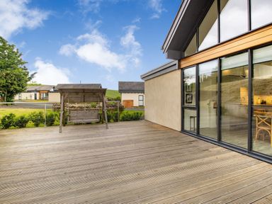 An outdoor wooden deck with a wooden swing and glass windows showing a kitchen inside at Forget Me Not Lodge in Bamburgh