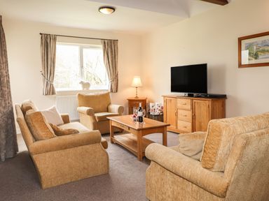 A living room with beige sofas a wooden coffee table a wooden TV cabinet a window with curtains and a small lamp at Tilly Mint Cottage Nr Ullswater