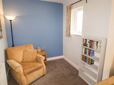 A small sitting area with an armchair next to a floor lamp and side table with a vase and a white bookshelf with books by a window at Tilly Mint Cottage Nr Ullswater