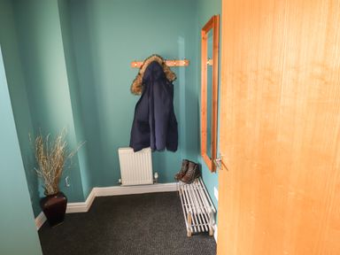 A hallway with a coat hanging on hooks a pair of boots on a rack a radiator a vase with branches and a wall mirror at Tilly Mint Cottage Nr Ullswater