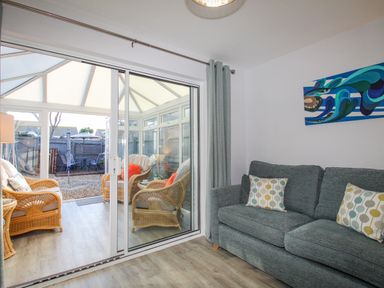 A living room with a gray sofa and patterned pillows next to a conservatory with wicker chairs and a garden outside at Chesil Retreat in Weymouth