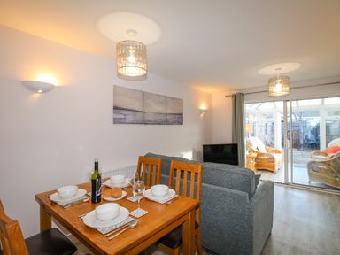 A dining area with a wooden table set for four next to a living room with a grey sofa and sliding glass doors leading to a room with wicker chairs at Chesil Retreat in Weymouth