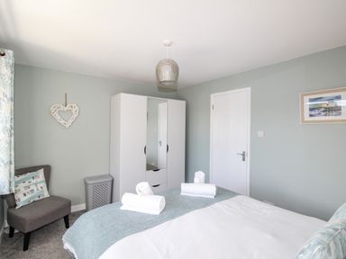 A bedroom with a bed, white wardrobe, grey chair with cushion, and a heart decoration on the wall at Chesil Retreat in Weymouth