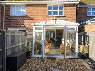A conservatory with wicker chairs and cushions facing a brick house with gravel ground and fenced garden at Chesil Retreat in Weymouth