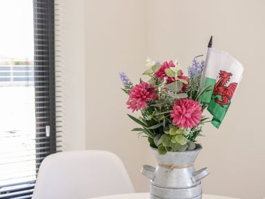 A metal vase with pink flowers and a small Welsh flag on a white table near a window with blinds at The Annexe in Llanfairpwllgwyngyll