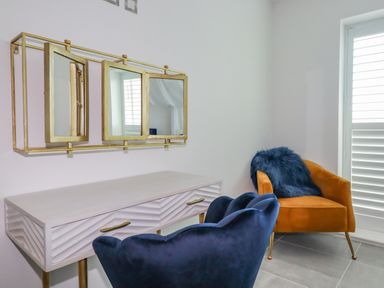 A room with a white desk and gold framed mirrors above it a blue chair in front of the desk and an orange chair with a blue cushion next to a window at 30 Cliff Edge in Newquay