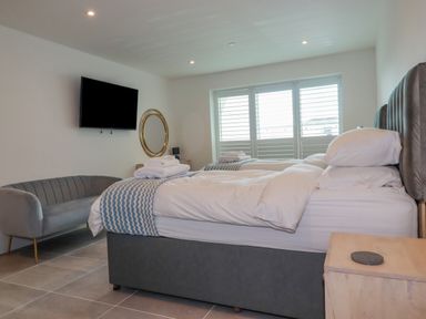 A bedroom with two beds a grey sofa a wall-mounted television and a window with shutters at 30 Cliff Edge in Newquay