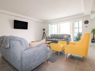 A living room with grey sofas yellow armchairs a wall-mounted TV and large windows at 30 Cliff Edge in Newquay