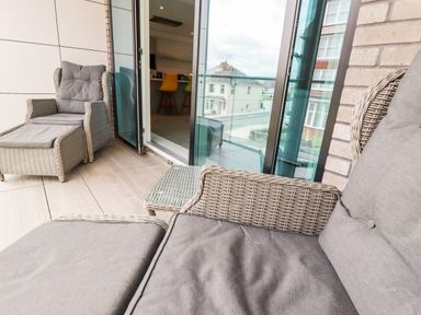 A balcony with wicker chairs and grey cushions and a glass door opening to a kitchen with bar stools at 30 Cliff Edge in Newquay