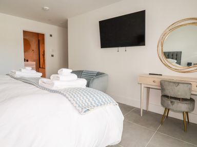 A bedroom with a bed, folded towels, a chair, a wall-mounted television, a desk with a round mirror, and a doorway to a bathroom at 30 Cliff Edge in Newquay