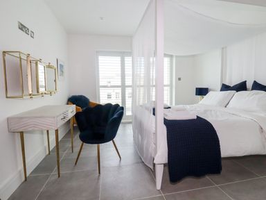 A bedroom with a canopy bed white bedding blue throw pillows and a blue chair next to a vanity table at 30 Cliff Edge in Newquay
