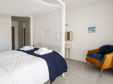 A bedroom with a four-poster bed with white and navy bedding towels on bed a mustard armchair with a blue throw a small vanity desk and cupboards at 30 Cliff Edge in Newquay