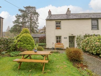 A garden with a wooden picnic table and bushes in front of a cream cottage with green door at Nook Cottage Rosthwaite near Keswick