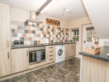 A kitchen with wooden cabinets a tiled backsplash an oven a washing machine a sink and a door at Nook Cottage Rosthwaite near Keswick