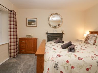 A bedroom with a wooden bed with floral bedding and rolled towels a wooden cabinet plaid curtains and a round wall mirror at Nook Cottage in Rosthwaite near Keswick