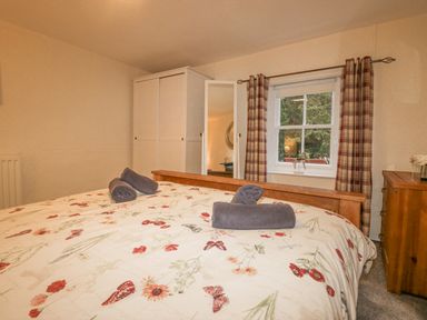 A bedroom with a bed covered in floral bedding with towels on top a wardrobe with a mirror and a window with plaid curtains at Nook Cottage in Rosthwaite near Keswick