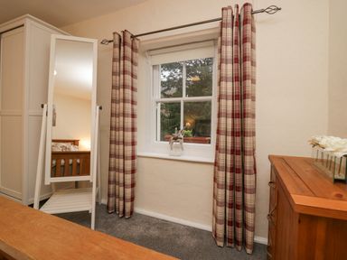 A bedroom window with plaid curtains a wooden chest of drawers a standing mirror and a wardrobe at Nook Cottage in Rosthwaite near Keswick