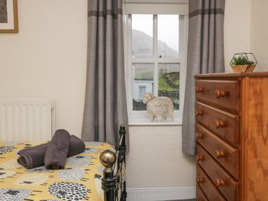 A bedroom with a metal bed frame and towels on the bed a wooden chest of drawers and a window with grey curtains and a decorative sheep on the sill at Nook Cottage in Rosthwaite near Keswick
