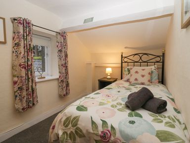 A small bedroom with a single bed with floral bedding a bedside table with a lamp and a window with floral curtains at Nook Cottage in Rosthwaite near Keswick