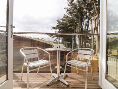 A balcony with two metal chairs and a round table with two mugs overlooking trees and a wooden fence at 11 Towan Valley in Porthtowan