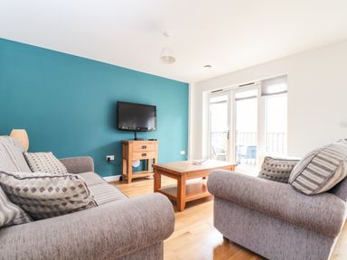 A living room with two gray sofas, a wooden coffee table, a wall-mounted TV, and glass doors leading to a balcony at 11 Towan Valley Porthtowan