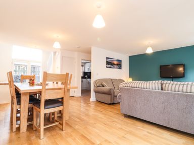 A dining area with wooden table and chairs next to a living room with sofas and a wall-mounted TV at 11 Towan Valley in Porthtowan