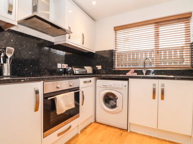 A kitchen with white cabinets a washing machine an oven and a window with blinds at 11 Towan Valley in Porthtowan
