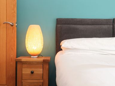 A bedside table with a lamp next to a bed with white bedding and a dark headboard at 11 Towan Valley in Porthtowan