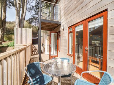 An outdoor patio with a metal table and blue chairs beside wooden house walls and glass doors at 11 Towan Valley in Porthtowan