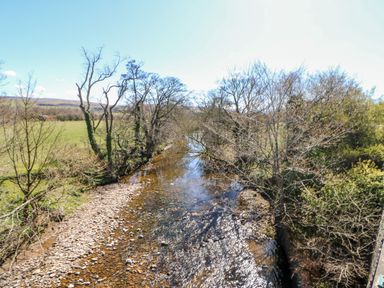 A shallow river with rocks on the banks and trees on both sides with fields and hills in the background at Summerseat in Kirkby Stephen