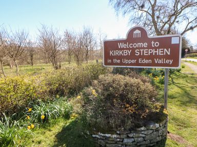 A welcome sign for Kirkby Stephen in the Upper Eden Valley beside a road with plants and trees in the background at Summerseat in Kirkby Stephen