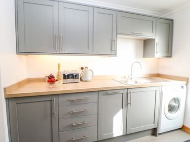 A kitchen with gray cabinets a wooden countertop a sink a washing machine a kettle a toaster and a bowl of fruit at Summerseat in Kirkby Stephen