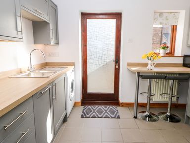 A kitchen with gray cabinets a sink a washing machine a wooden door a small dining area with two stools and flowers at Summerseat in Kirkby Stephen