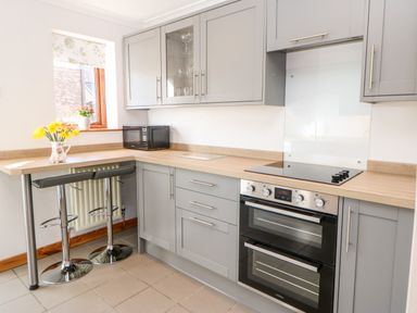 A kitchen with gray cabinets a built-in oven a microwave two bar stools and a vase with flowers on the counter at Summerseat in Kirkby Stephen