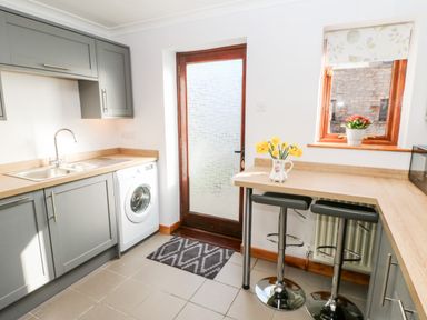 A kitchen with grey cabinets a washing machine a wooden door and a small breakfast bar with two stools and flowers at Summerseat in Kirkby Stephen
