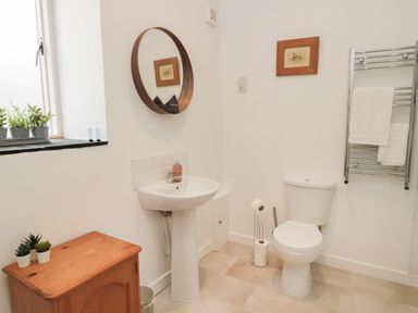A bathroom with a pedestal sink and round mirror above it a toilet with a toilet roll holder a heated towel rail with towels a wooden cabinet with plants on top and a small window at The Bakery in Middleton near Belford