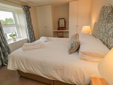 A bedroom with a bed featuring patterned headboard and cushions two folded towels on the bed bedside lamps window with patterned curtains and a small plant on the windowsill built-in white wardrobes and a wooden dressing table with a mirror and chair at 46 Castle Street in Norham