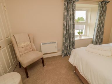 A bedroom corner with a beige chair with a blanket a window with curtains and a bed with folded towels at 46 Castle Street in Norham
