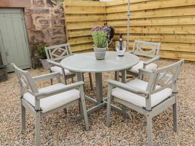An outdoor patio area with a round table and four chairs with cushions a potted plant a bottle and two glasses on the table and a barbecue against a wooden fence at 46 Castle Street in Norham