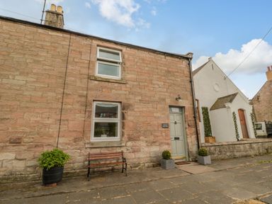 Exterior view of a stone house with two windows a door two potted plants and a bench on the sidewalk at 46 Castle Street in Norham