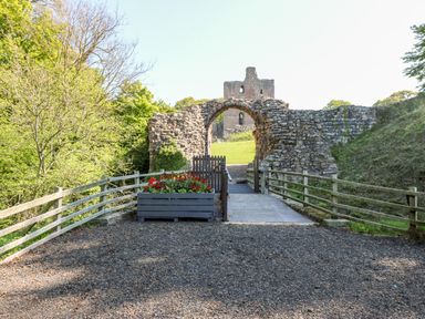A stone archway with a wooden gate and flower planter on a gravel path near castle ruins at 46 Castle Street in Norham