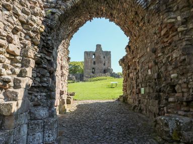 View of a stone archway leading to a grassy area with the ruins of a stone building in the background at 46 Castle Street in Norham