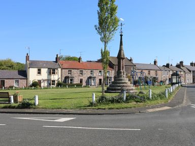 A grassy area with a stone monument and swings next to a row of houses at 46 Castle Street in Norham