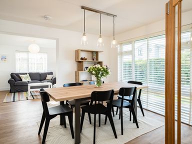 A dining area with a wooden table and six black chairs a vase with flowers on the table a shelving unit on the wall and a living room with a gray sofa and white coffee table visible through an open doorway at Obo's Retreat in St Minver near Rock