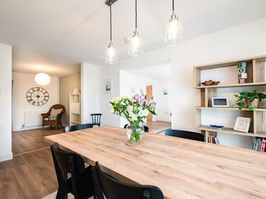 A dining area with a wooden table, black chairs, flowers in a vase, pendant lights, wall shelves, and a chair in an adjoining room at Obos Retreat in St Minver near Rock