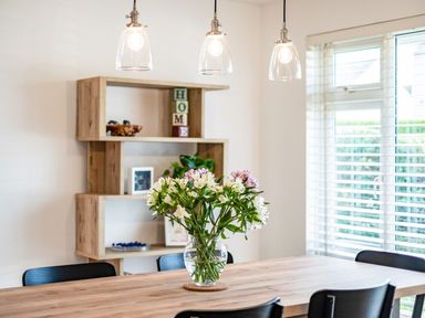 A dining table with a glass vase of flowers and black chairs near a wooden shelving unit and window blinds at Obo's Retreat in St Minver near Rock